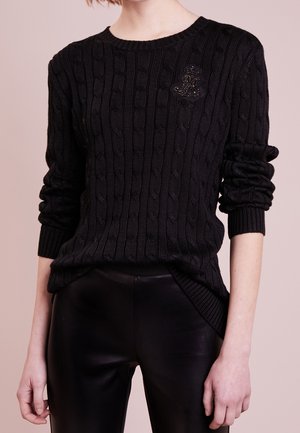 Nainen pukeutunut mustaan palmikkoneulepaitaan, jossa on kirjailtu yksityiskohta rinnassa, sekä mustiin nahkahousuihin, seisoo vaaleaa taustaa vasten.