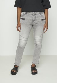 Jeans de ganga cinza com um corte justo, apresentando costuras visíveis, bolsos laterais com zíper e detalhes em painéis nos joelhos, combinados com sandálias pretas.
