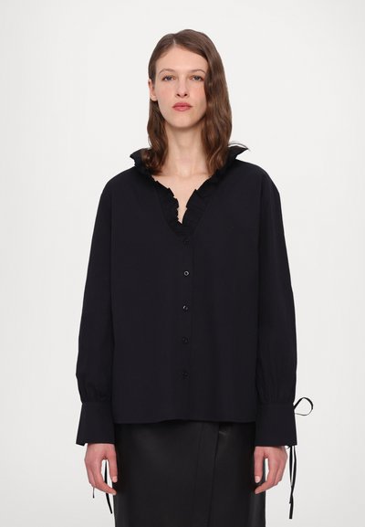 Chemise noire à boutons avec un col à volants, des manches longues et des liens aux poignets. Fabriquée dans un tissu à la texture douce.