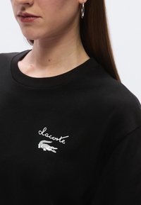 Čierne bavlnené tričko s okrúhlym výstrihom, na ktorom je biely nápis "Lacoste" a vyšívané logo krokodíla na ľavej strane hrudníka.