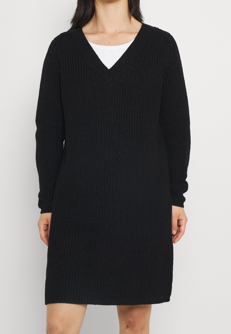 Svart stickad klänning med V-ringning, långa ärmar och en lös passform. Texturen är ribbad, och fållen slutar ovanför knäet.