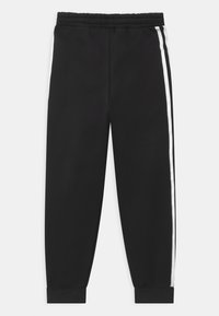 Černé sportovní kalhoty z hladké tkaniny, se širokými bílými pruhy po stranách, elastickým pasem a zúženými manžetami na kotnících.