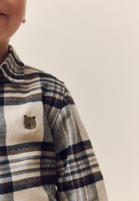 Kariertes Flanellhemd in Beige, Blau und Braun mit einer Bärenstickerei auf der Brusttasche. Verfügt über einen spitzen Kragen und einen Knopfverschluss.