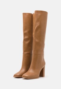 Bottes en cuir marron, hautes, avec une texture lisse, un bout carré et un talon carré. Présente des lignes épurées et un design minimaliste.