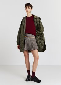 Olivgrüne gesteppten Jacke, bordeauxfarbenem Pullover, grauer karierten gefalteten Rock, schwarzen Schuhen und bordeauxfarbenen Socken. Lässiges, geschichtetes Outfit mit verschiedenen Texturen.