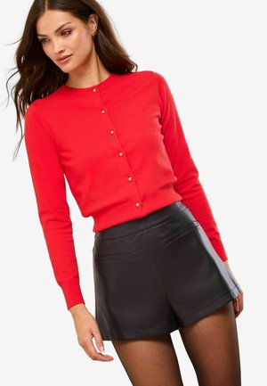 Frau in einer figurbetonten roten Knopfkardi mit goldenen Knöpfen und schwarzen Leder-Shorts, die vor einem schlichten weißen Hintergrund posiert.