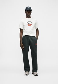 Άνδρας που φοράει λευκό μπλουζάκι με το λογότυπο της Nike, μαύρη φόρμα Nike, μπεζ αθλητικά παπούτσια και μαύρο καπέλο Nike, στέκεται μπροστά σε απλό ανοιχτό γκρι φόντο.