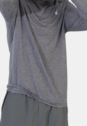 Mężczyzna w szarej, teksturowanej koszuli z długim rękawem i małym logo pająka na piersi oraz ciemnoszarych spodniach, poprawiający rękawy.