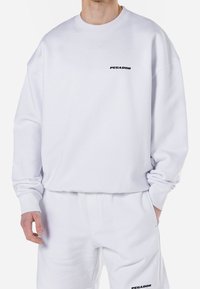 White sweatshirt made of cotton blend, featuring ribbed cuffs and a crew neck. Black "PEGADOR" logo on chest. Smooth texture and relaxed fit.