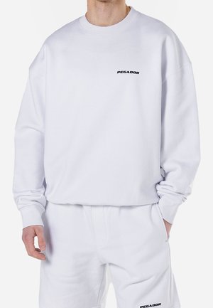 White sweatshirt made of cotton blend, featuring ribbed cuffs and a crew neck. Black "PEGADOR" logo on chest. Smooth texture and relaxed fit.