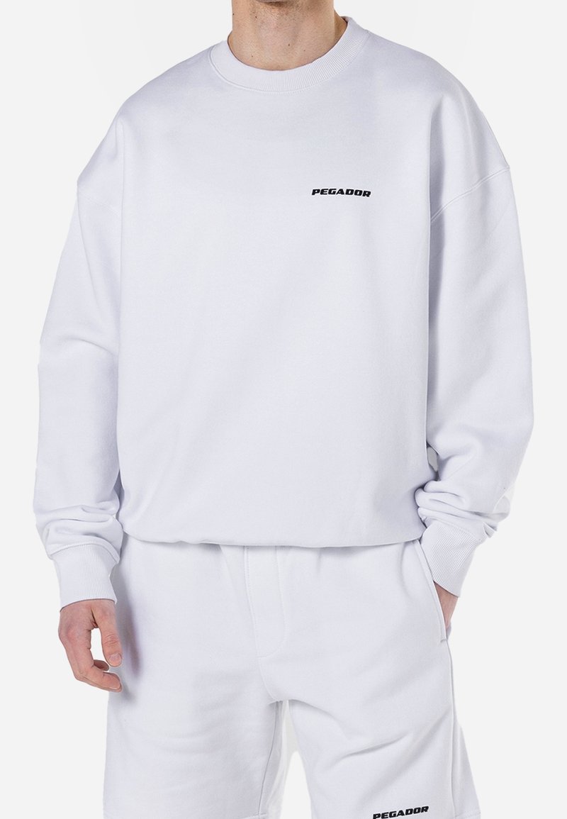 White sweatshirt made of cotton blend, featuring ribbed cuffs and a crew neck. Black "PEGADOR" logo on chest. Smooth texture and relaxed fit.