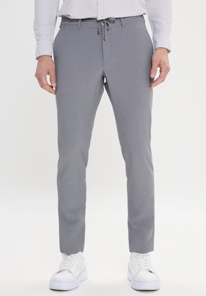 Hombre con pantalones ajustados grises con cintura de cordón y zapatillas blancas, de pie frente a un fondo liso.