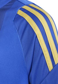 Sininen urheilu-paita tekstuuripinnalla, jossa on kolme näkyvää keltaista raitaa olkapäällä ja erottuvaa sinistä paneelointia.