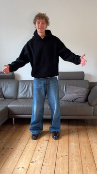 Juoda laisvo kirpimo hoodie, šviesiai mėlyni laisvi džinsai, blizgūs juodi bateliai. Stovi ant medinių grindų prie pilko sofos su raštuotomis pagalvėlėmis.