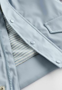 Veste imperméable bleu clair avec une fermeture éclair, des boutons-pression et une doublure intérieure rayée. Présente une texture lisse et un design simple.