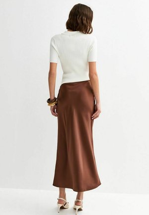 Kobieta o brązowych włosach do ramion stoi tyłem, ubrana w biały prążkowany top z krótkim rękawem, brązową satynową spódnicę i beżowe szpilki.