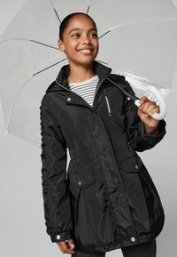 Giacca antivento nera e leggera con tasca con zipper, cinture elastiche ai lati e due tasche frontali. Il modello tiene un ombrello trasparente. Camicia a righe sotto.