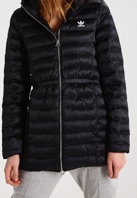 Svart pufferjacka med dragkedja och dragsko i midjan. Har en huva och en liten vit Adidas-logotyp på bröstet, quiltad textur.