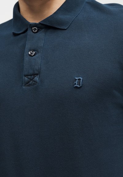 Σκούρο μπλε πόλο μπλούζα με δύο κουμπιά και ένα μικρό κεντημένο γράμμα "D" στην αριστερή πλευρά του στήθους.
