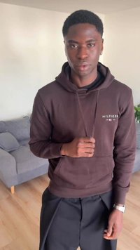 Bărbat tânăr îmbrăcat cu hanorac Tommy Hilfiger maro închis și pantaloni negri, stând în interior lângă o canapea gri cu podea din lemn deschis la culoare.