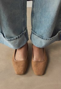 Brune suedestøvler med firkantet tådesign, placeret mellem oprullede lyseblå denimjeans på en neutral baggrund.