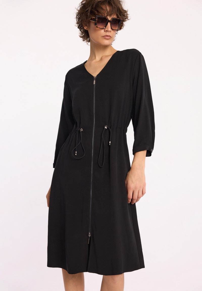 Czarna sukienka wykonana z gładkiego materiału, z dekoltem w serek, suwakiem z przodu, rękawami o długości trzech czwartych, talią regulowaną sznurkiem oraz srebrnymi detalami.