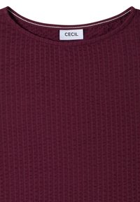 Gros plan d'un haut en tissu côtelé bordeaux avec un col rond et une étiquette blanche portant la mention "CECIL" cousue à l'intérieur du col.