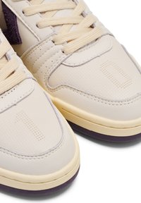 Beige Leder-Sneaker mit strukturiertem Obermaterial, hellen Schnürsenkeln und lila Akzenten an der Sohle. Verfügt über Perforationen und ein numerisches Designelement.
