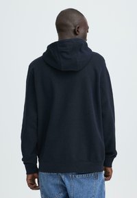 Marineblauwe capuchontrui met een gestructureerde stof, geribbelde manchetten en zoom. Gedragen met lichtblauwe denim shorts, rugaanzicht.