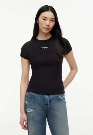 Joven mujer con cabello largo y oscuro que lleva una camiseta negra ajustada con el texto "tommy jeans" y vaqueros azul desgarrados, posando sobre un fondo blanco.