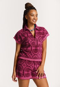 Růžový textilní set obsahující top s krátkým rukávem a límečkem a odpovídající šortky, obě s mušlovými a hvězdicovými vzory a třásněmi na okrajích.