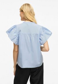 Gaiši zila blūze ar puff piedurknēm, savelkamu aizmugures detaļu un apaļu kakla izgriezumu, izgatavota no gludas auduma, kopā ar melniem biksēm.