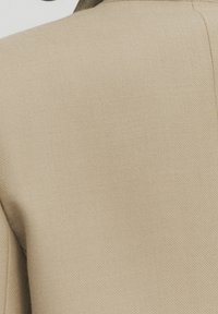 Tessuto beige con una superficie texture, forma strutturata con colletto e linee pulite. I dettagli delle cuciture sono visibili lungo i bordi.