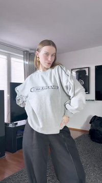 Svetlo šedý oversized mikina s čiernym potlačeným nápisom "LET'S FRANS". Má spustené ramená a priliehavé manžety. Skombinovaná s voľnými čiernymi nohavicami.