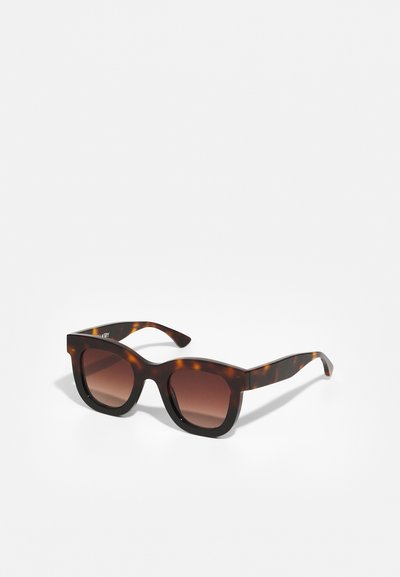 Lunettes de soleil en écaille de tortue avec une monture épaisse et angulaire. Verres dégradés marron et branches légèrement épaisses. Finition en plastique lisse avec une texture brillante.