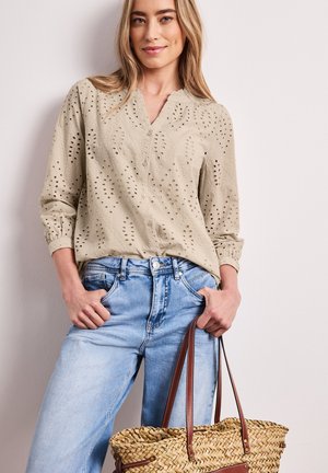 Femme portant un chemisier beige à œillets et un jean bleu clair, tenant un sac fourre-tout en paille tressée avec des poignées en cuir marron.