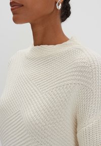 Maglione lavorato a maglia di colore crema con un motivo testurizzato, caratterizzato da un collo alto e un dettaglio a punto diagonale su un lato.