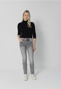 Maglione a collo alto nero, jeans slim-fit grigi, scarpe da ginnastica bianche. Modello in piedi davanti a uno sfondo neutro, mani nelle tasche. Look casual.