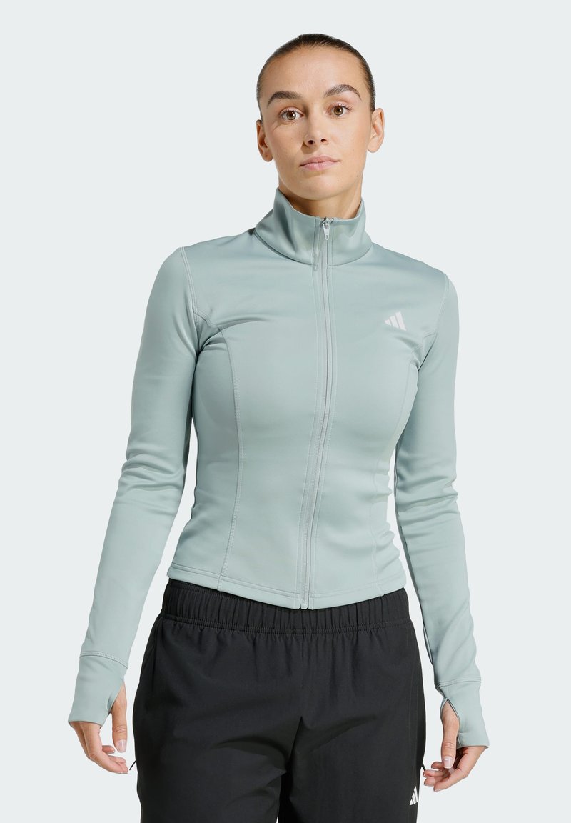 Γυναίκα που φοράει ανοιχτό μπλε αθλητικό μπουφάν με φερμουάρ και τρύπες για τους αντίχειρες, καθώς και μαύρο αθλητικό παντελόνι, στέκεται με το σώμα της προς τα εμπρός πάνω σε ένα απλό φόντο.