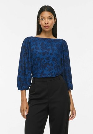 Femeie cu păr lung și închis la culoare, care poartă o bluză albastră cu model floral, mâneci bufante trei-sferturi și pantaloni negri cu talie înaltă pe un fundal alb.