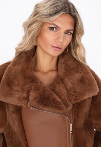 Blonde vrouw met golvend haar draagt gouden oorbellen en een bruin faux fur en leren jasje met een opvallende rits.