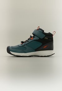 Παπούτσι πεζοπορίας μεσαίου ύψους σε τυρκουάζ και μαύρο, με ιμάντα Velcro, παχύ λαστιχένιο πέλμα και πορτοκαλί λεπτομέρειες με υφή στην επιφάνεια.