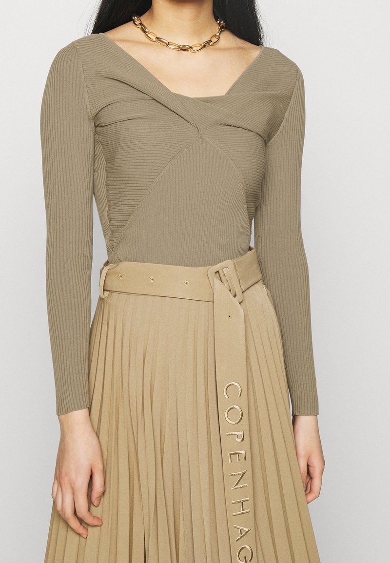 Femme portant un haut taupe côtelé à manches longues avec un décolleté unique et une jupe plissée beige taille haute avec ceinture assortie et collier chaîne doré.