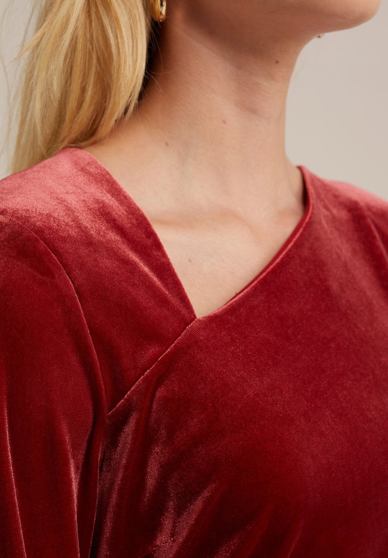 Czerwony aksamitny top z głębokim dekoltem w kształcie V, gładką teksturą i kanciastym szwem ramiennym, podkreślający bogaty kolor i luksusowy materiał.