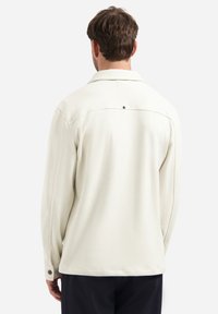 Man met kort bruin haar, gekleed in een off-white shirt met lange mouwen en structuur, gezien van achteren tegen een effen achtergrond.