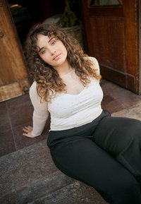 Langärmliges durchsichtiges Oberteil mit silbernen Punkten, kombiniert mit hoch taillierten schwarzen Hosen. Lockiges Haar und dezentes Make-up, sitzend auf steinernen Treppen.