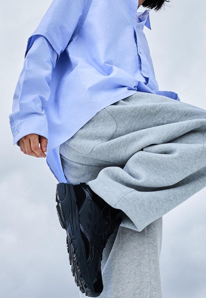 Svijetloplava košulja s dugim rukavima, sivi trenirke i crne tenisice. Košulja ima opušteni kroj, a tenisice imaju teksturiranu potplat.