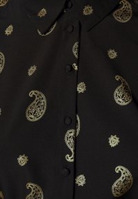 Musta pusero, jossa on kultainen paisley-kuvio, varustettuna kauluskannalla ja nappilistalla edessä. Pehmeä kangas, jossa on sileä tekstuuri.