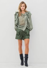 Grönmönstrad blus med volanger, kombinerad med mörkgröna shorts. Bärs med knähöga svarta stövlar. Enfärgad bakgrund.