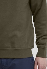 Felpa verde oliva con una texture morbida, polsini e orlo a coste. Include un piccolo etichetta tessuta sul lato. Abbinata a pantaloni blu navy.